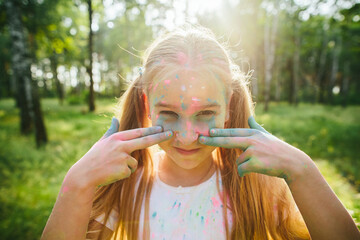 Caucasian girl with colorful powder playfully points to her eyes in forest light. Fun childhood moment, emotion, outdoor freedom and authentic personal expression.
