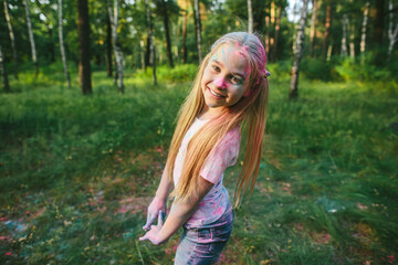 Joyful young girl with long hair and pigtails smiling at camera after throwing colorful powder...