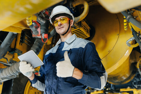 Worker mechanic with tablet computer under mining truck in garage, service center of industrial machinery
