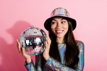 Stylish young woman holding a disco ball against a pink background in a shiny outfit and...