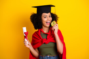 Confident young graduate holding diploma and award medal wearing casual cap and vibrant cape on yellow background