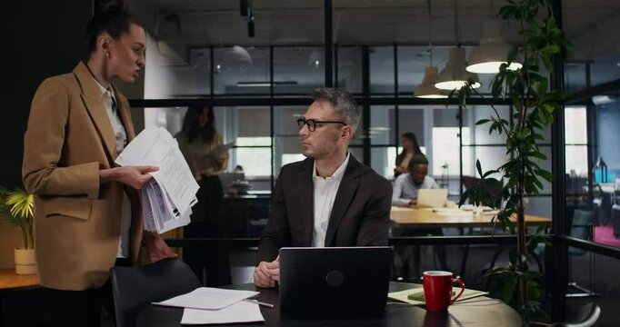 The boss shouts at his subordinate and throws documents at him. A man is sitting at a table in front of an open laptop with a puzzled look