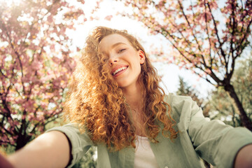 Joyful young woman with curly red hair enjoying a sunny day outdoors by blooming trees in a...