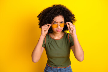 Confident young woman adjusting stylish yellow sunglasses against a vibrant yellow background with...