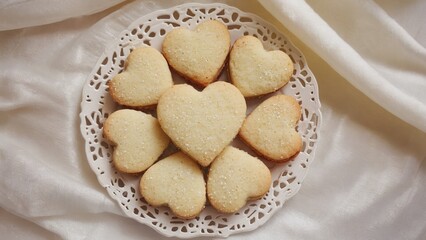 A plate of heart-shaped cookies arranged neatly on a lace doily. The cookies are light golden in color, symbolizing love and celebration during holidays.