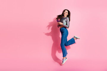 Young fashion-forward woman joyfully jumping against a pink background holding a smartphone while...