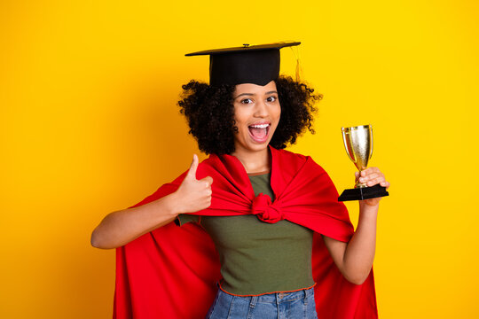 Young woman celebrating achievement wearing graduation cap and holding a trophy against a vibrant yellow background - Powered by Adobe