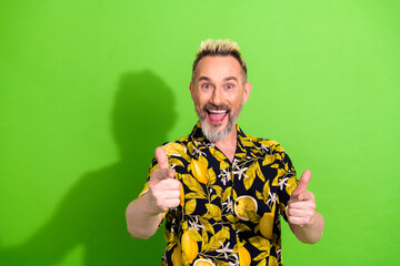 Charismatic man in a vibrant summer shirt giving enthusiastic thumbs up gesture against a green...