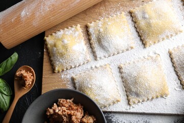 Uncooked ravioli, rolling pin, bolognese and basil on black table, flat lay