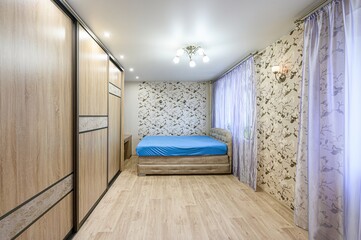Fototapeta premium cozy bedroom features a large wooden wardrobe, a bed with a blue sheet, patterned wallpaper, and soft violet curtains