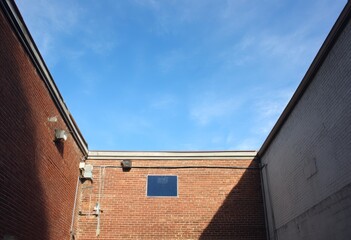Low-angle view of a brick-walled alleyway, a small square window punctuates a brick wall under a vibrant blue sky