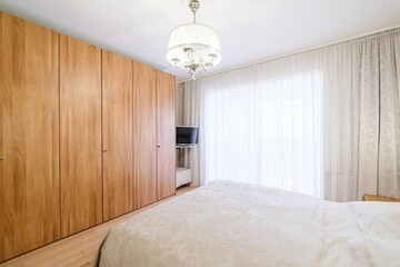 bright bedroom features a bed with patterned bedding, a large wooden closet, and a chandelier....
