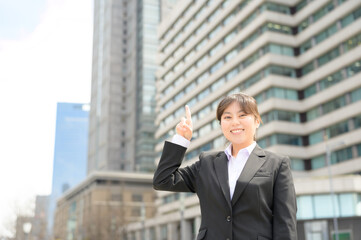 春のオフィス街で明るく手を挙げるスーツ姿の新入社員の女性
