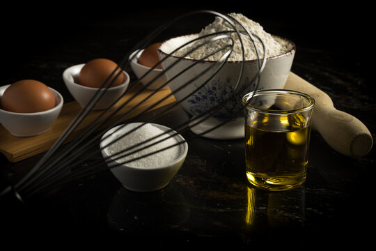 Ingredientes para hornear, incluyendo huevos, harina, az&uacute;car y aceite, dispuestos con utensilios de cocina sobre un fondo oscuro. 