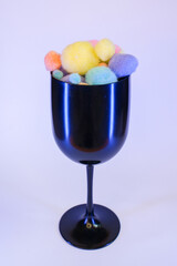 A black wine glass filled with colorful soft pom poms on a clean white background. Minimalistic and playful composition.
