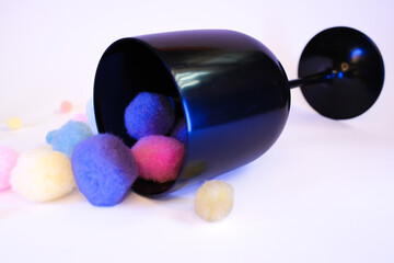 Minimalist and playful still life with a black wine glass tipped over, spilling soft colorful pom...