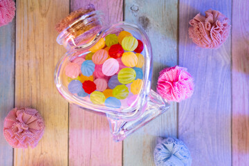 Colorful pastel candies in a heart-shaped glass jar, surrounded by decorative pom-poms on a wooden...