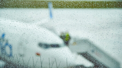 Rain falling on airport window blurs airplane and ground crew preparing for departure on a rainy day