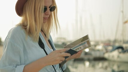 Blonde traveler wearing sunglasses and straw hat reviewing guidebook near marina boats, embodying relaxed vacation planning mood. Europe travel. Lifestyle, vacation, tourism, active life