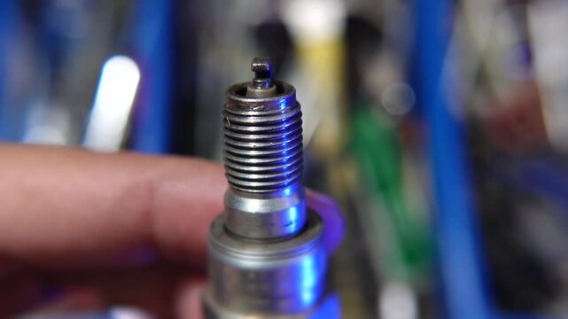 A detailed close-up image featuring a hand holding a worn spark plug against a blurred background of mechanical tools and equipment.