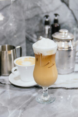 A glass of coffee with whipped cream on top sits on a table next to a cup