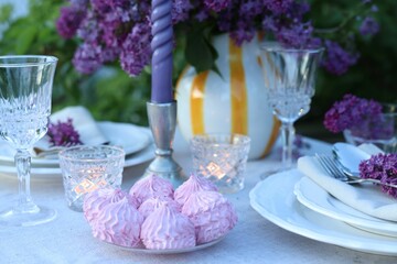 Beautiful lilac flowers, zephyrs, plates, cutlery, glasses and candles on table in garden, closeup