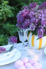 Beautiful lilac flowers, zephyrs, plates, cutlery, glasses and burning candles on table in garden, closeup