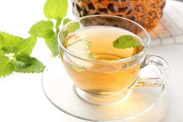 Aromatic lemon balm tea in glass cup, teapot and fresh leaves on white table, closeup