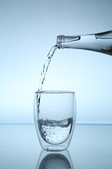 Pouring water from bottle into glass at mirror table against light blue gradient background, closeup