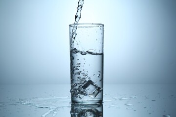Pouring water into glass at mirror table against light grey gradient background, closeup