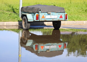 A two-wheeled outdoor car trailer covered with a tarp is reflected in a puddle, Dybenko Street, St. Petersburg, Russia, June 20, 2025
