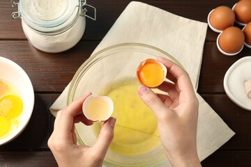 Woman separating egg yolk from white over glass bowl at wooden table, top view