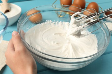 Woman whisking cream with hand mixer at light blue wooden table, closeup