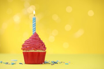 Burning candle in cupcake and sprinkles on yellow background with blurred lights, space for text