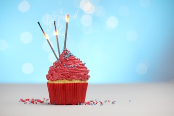 Burning sparklers in cupcake and sprinkles on white table against light blue background with...