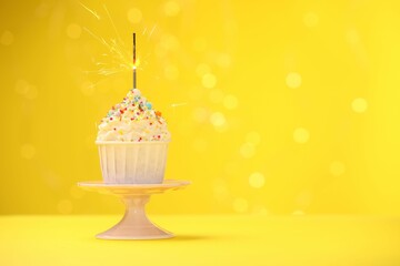 Burning sparkler in cupcake, sprinkles and stand on yellow background with blurred lights, space...