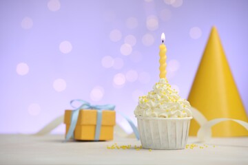 Burning candle in cupcake, sprinkles and party decor on white table against violet background with blurred lights, space for text