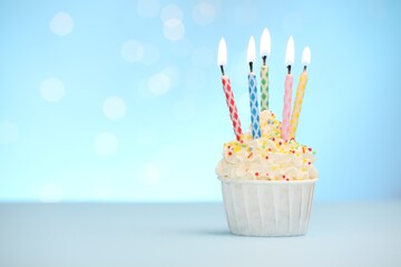 Burning candles in cupcake and sprinkles on white table against light blue background with blurred...