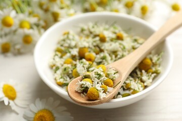 Dry chamomile flowers in bowl and spoon on light table, closeup