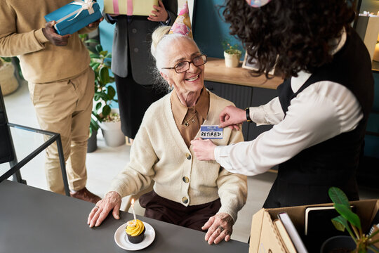 Senior Caucasian woman sitting at desk smiling while female colleague attaching retirement badge to her chest during office celebration
