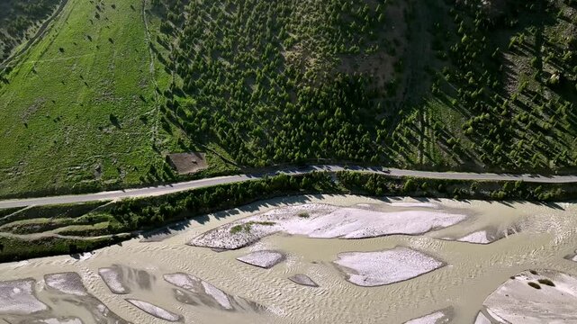 4K Aerial side tracking drone video shot of moving vehicles driving along a winding mountain road beside flowing river. Scenic natural terrain, sharp curves, and dramatic elevation, pine tree shadows.