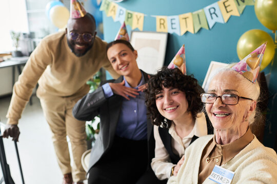 Group of diverse adults and senior Caucasian woman wearing party hats celebrating retirement in studio, senior woman smiling and taking selfie while others posing and looking cheerful