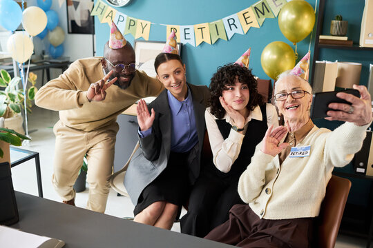 Group of senior Caucasian woman, middle aged Caucasian woman, middle aged Black man, and young adult Caucasian woman smiling and waving while taking selfie during retirement celebration