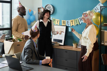 Senior Caucasian woman standing and smiling while talking with middle aged Black man and two colleagues wearing party hats celebrating retirement in office setting