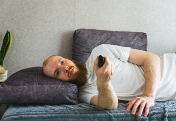 Unmotivated bearded man lying in bed with remote control, looking bored and uninterested. Concept...