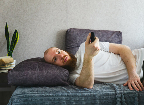 Unmotivated bearded man lying in bed with remote control, looking bored and uninterested. Concept of lazy day, boredom, or passive leisure at home.

