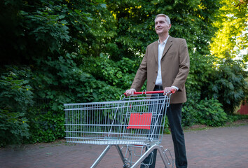 Man in brown blazer pushing empty shopping cart outdoors in park setting.