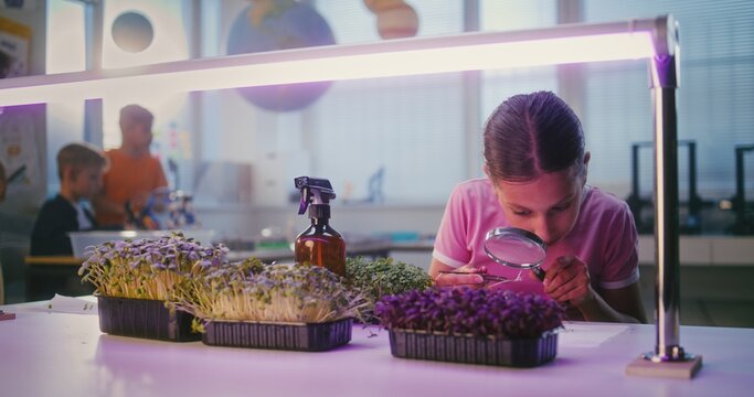Talented Girl Sitting at Table and Using Smartphone, Conducting Biology Experiment on Plants with Tweezer and Magnifying Glass. Primary School Children During Lesson in Classroom. STEM Education.