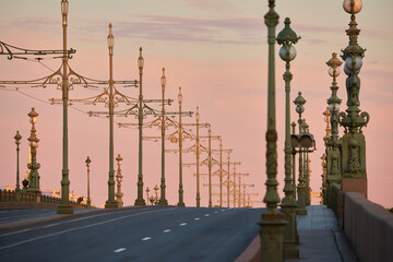 rows of lampposts and poles with electric grids for a tram on an empty Troitskiy Bridge at a pink sunset, nobody