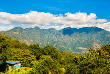 Scenic view of a lush forest and calm lake in Guatemala, with a green volcanic hill rising near the shoreline and vast mountains fading into the misty background under soft clouds.